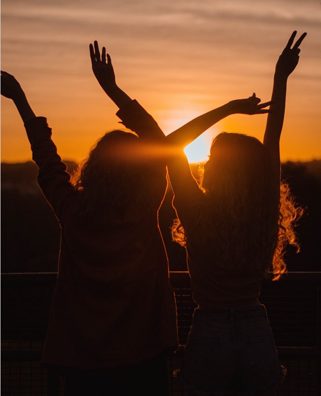 Dos chicas de espaldas con los brazos levantados en una puesta de sol.
