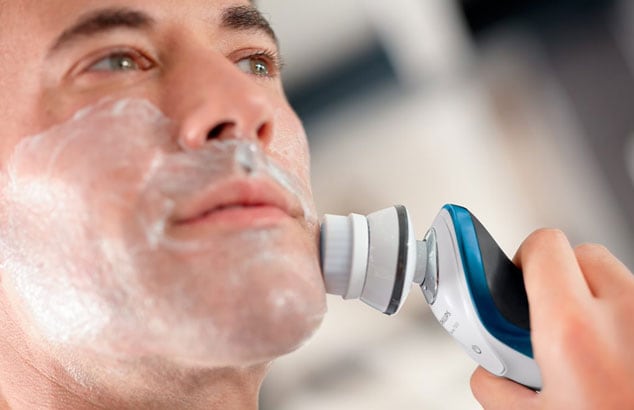Primer plano de la cara de un hombre con espuma de afeitar utilizando una maquinilla eléctrica en la mejilla