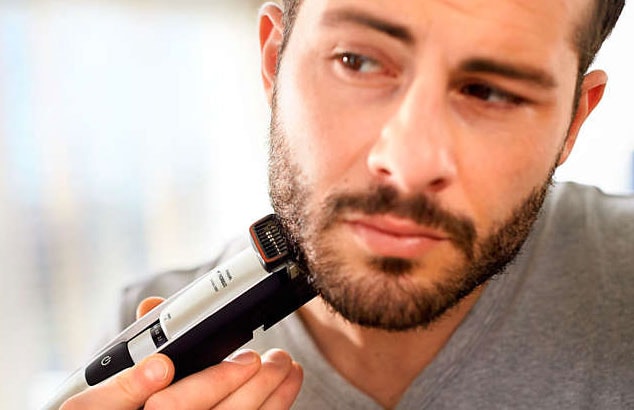 Hombre moreno y con barba, con una camiseta gris, utilizando una maquinilla de afeitar eléctrica en la línea de la mandíbula