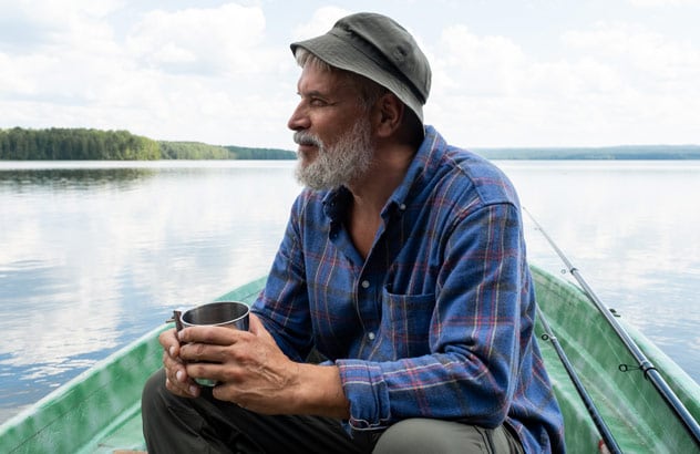 Un hombre mayor con barba gris, sombrero de pescador y camisa azul de cuadros sentado en un barco y mirando a lo lejos