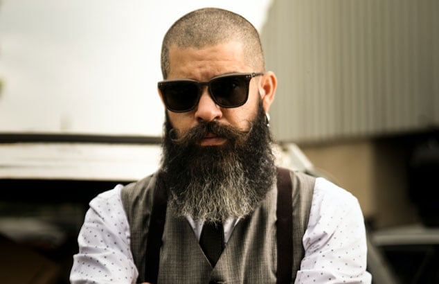 Foto de un hombre con el pelo corto, barba larga y completa, gafas de sol y tatuajes en los antebrazos