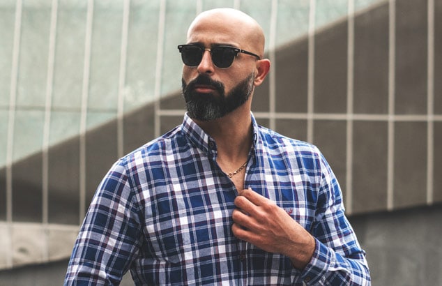 Un hombre elegante y calvo con barba bien recortada, camisa azul de cuadros y gafas de sol 