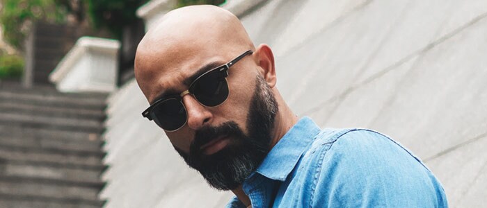 Un hombre calvo con barba oscura ligeramente canosa, gafas de sol y camisa vaquera 