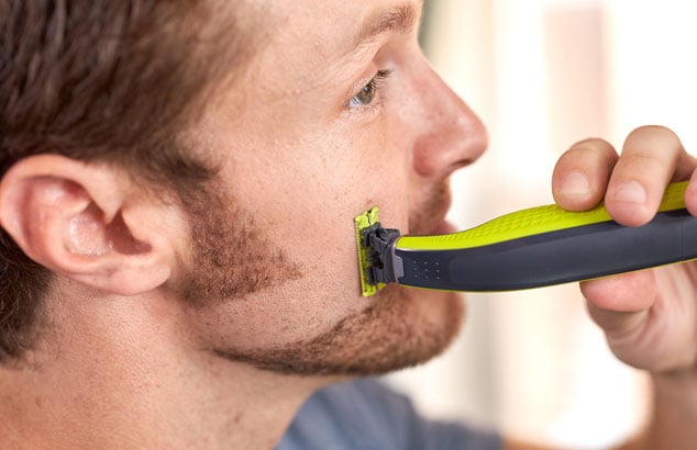 Un hombre afeitándose parte de su barba, dando forma a unas patillas que conectan con su barba