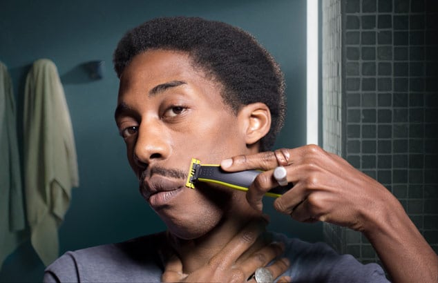 Un joven de piel oscura recortándose el bigote con Philips OneBlade frente a un espejo en un baño