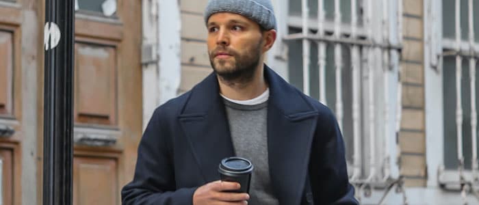 Un hombre con barba de tres días, gorro y abrigo, caminando por una calle residencial con una taza de café