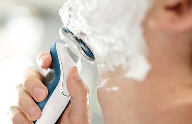 Primer plano de la barbilla de un hombre desenfocado con espuma de afeitar y una maquinilla eléctrica blanca y azul